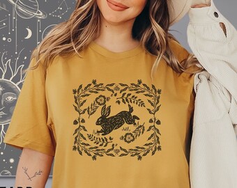 May include: A mustard yellow t-shirt with a black and white illustration of a rabbit running through a floral wreath. The text "MUSTARD" is visible at the bottom of the image.