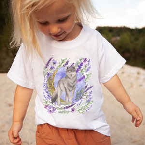 May include: White t-shirt with a watercolor illustration of a wolf standing in front of a crescent moon surrounded by lavender flowers.