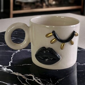 Könnte beinhalten: Eine weiße Keramiktasse mit einem schwarzen und goldenen Auge und einem schwarzen Lippen-Design. Die Tasse hat einen abgerundeten Griff.