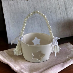 May include: A white ceramic candle holder with star cutouts and a beaded handle. The handle is made of white pearls. White tassels and ribbons are attached to the sides. A lit candle is in the background.