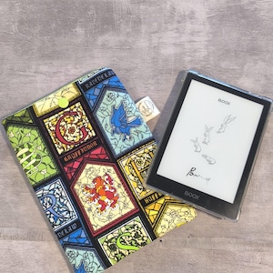 Pode incluir: Uma capa de tecido com um padrão de vitral colorido com brasões e texto incluindo Gryffindor, Ravenclaw e Hufflepuff, ao lado de um leitor eletrônico Boox preto exibindo um gráfico de quatro coelhos e o texto Power of.