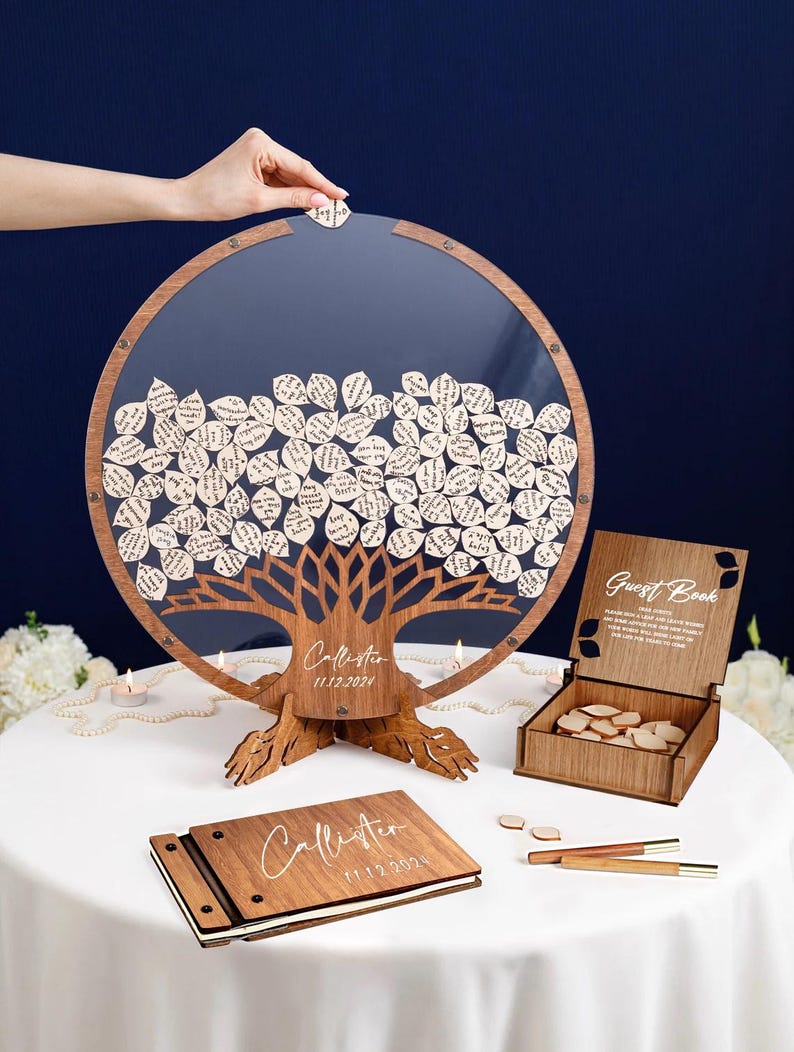 May include: A wooden guest book alternative with a tree design. The tree is made of wood and has leaves that can be written on. The leaves are placed in a clear circular frame. The guest book also includes a wooden box with a lid and a small wooden book. The box and book are both engraved with the text "Guest Book".