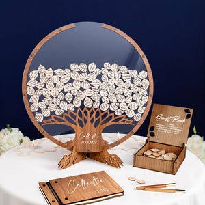 May include: A wooden guest book with a tree design and a clear acrylic cover. The tree is filled with small wooden leaves with handwritten messages. The guest book is on a white table with a wooden box and two pens.