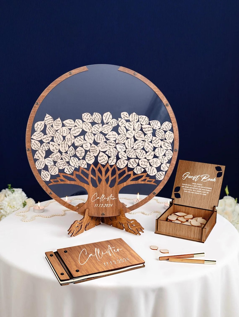 May include: A wooden guest book with a tree design and a clear acrylic cover. The tree is filled with small wooden leaves with handwritten messages. The guest book is on a white table with a wooden box and two pens.