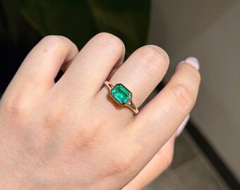 以下が含まれることがあります： ベゼルセッティングで留められた長方形のエメラルドカットの緑色の宝石が特徴のゴールドリング。バンドは滑らかで磨き上げられており、鮮やかな緑色と中央の石のすっきりとしたラインを際立たせるシンプルなデザインです。