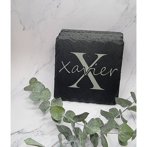 May include: Set of four black slate coasters with the name "Xavier" engraved in white. The coasters are stacked on top of each other and are sitting on a white marble surface.