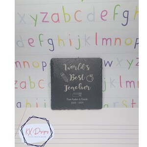 May include: A black slate coaster with the text "World's Best Teacher" engraved in silver. The coaster also includes a pencil, apple, and book icon. The text "From Kaden & Klarah. 2023-2024" is also engraved on the coaster.