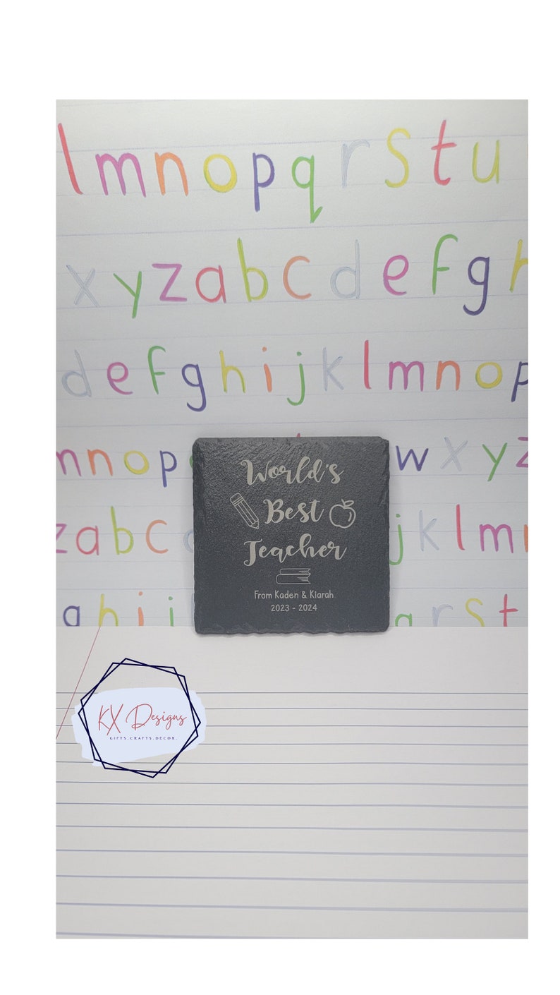 May include: A black slate coaster with the text "World's Best Teacher" engraved in silver. The coaster also includes a pencil, apple, and book icon. The text "From Kaden & Klarah. 2023-2024" is also engraved on the coaster.
