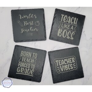 May include: Four black slate coasters with white text. The coasters feature teacher-themed sayings: "World's Best Teacher", "Teach Like A Boss", "Born To Teach Forced To Grade", and "Teacher Vibes".