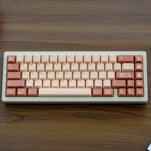 May include: A light beige and brown mechanical keyboard with a light beige space bar and brown keys. The keys have various symbols and icons on them, including a cat, a coffee cup, and a pencil.