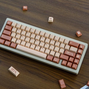 May include: A light green mechanical keyboard with a beige and brown keycap set. The keycaps feature various symbols and icons, including a cat, a coffee cup, and a feather. The keyboard is on a wooden surface.