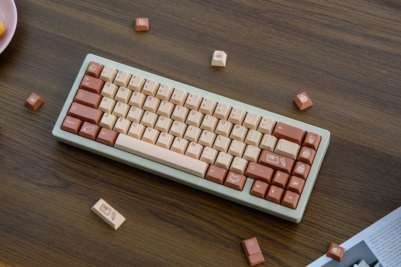May include: A light green mechanical keyboard with a beige and brown keycap set. The keycaps feature various symbols and icons, including a cat, a coffee cup, and a feather. The keyboard is on a wooden surface.