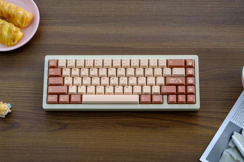 May include: A light beige and brown mechanical keyboard with a light beige space bar and brown keys. The keys have various symbols and icons on them, including a cat, a coffee cup, and a pencil.