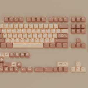 May include: A full keyboard set with beige and brown keycaps. The keycaps feature various symbols and icons, including a coffee cup, a cat, a cake, and a camera. The keyboard is laid out in a standard QWERTY configuration.