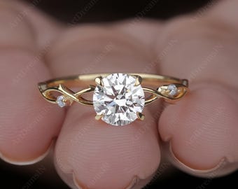 Könnte beinhalten: Ein goldener Verlobungsring mit einem großen, runden, klaren Edelstein als Mittelpunkt. Das Band hat ein zartes, ineinander verschlungenes Design mit kleinen Akzentsteinen auf beiden Seiten des Hauptsteins. Der Ring wird in einer Hand gehalten.