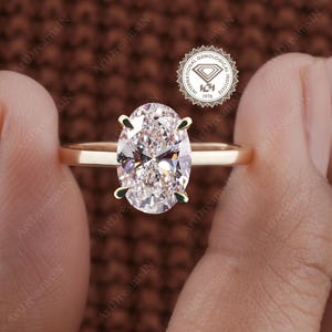 May include: A gold engagement ring with an oval-cut diamond. The diamond is secured by four prongs. The ring is held between two hands, with the International Gemological Institute logo visible in the background.