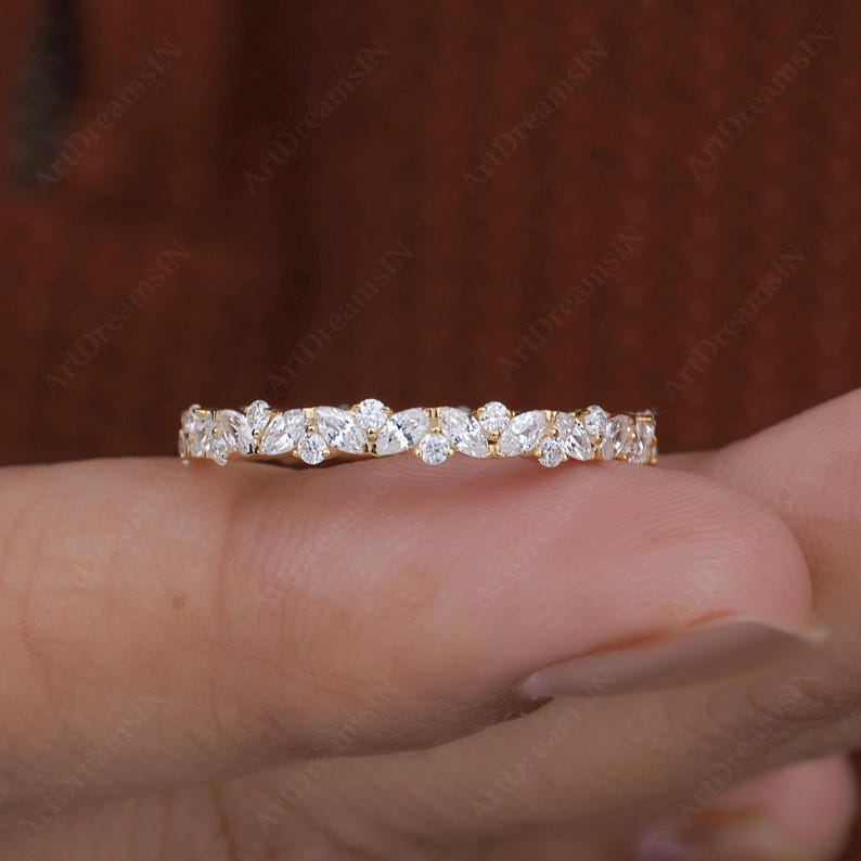 May include: A delicate gold ring featuring a continuous pattern of marquise and round-cut gemstones. The ring's design showcases alternating shapes, creating a sparkling, elegant band. The ring is held against a blurred background.