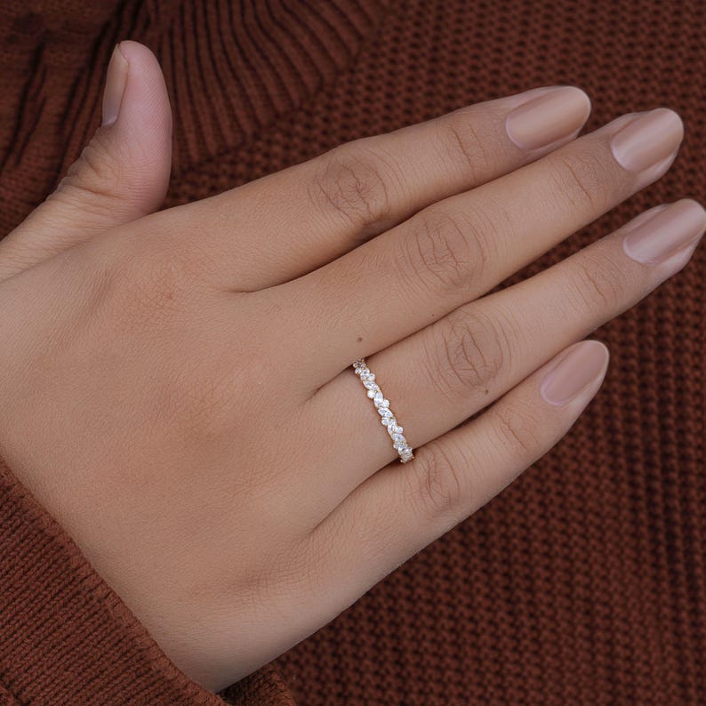 May include: A delicate, silver-toned ring with a continuous pattern of small, clear gemstones. The ring is shown on a hand with neutral-toned nail polish, set against a warm brown background. The ring's design is simple and elegant.