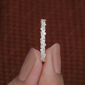 May include: A gold band ring with a continuous pattern of white gemstones. The ring is held by a person's hand against a dark brown background. The ring's gemstones are pear and round shaped.