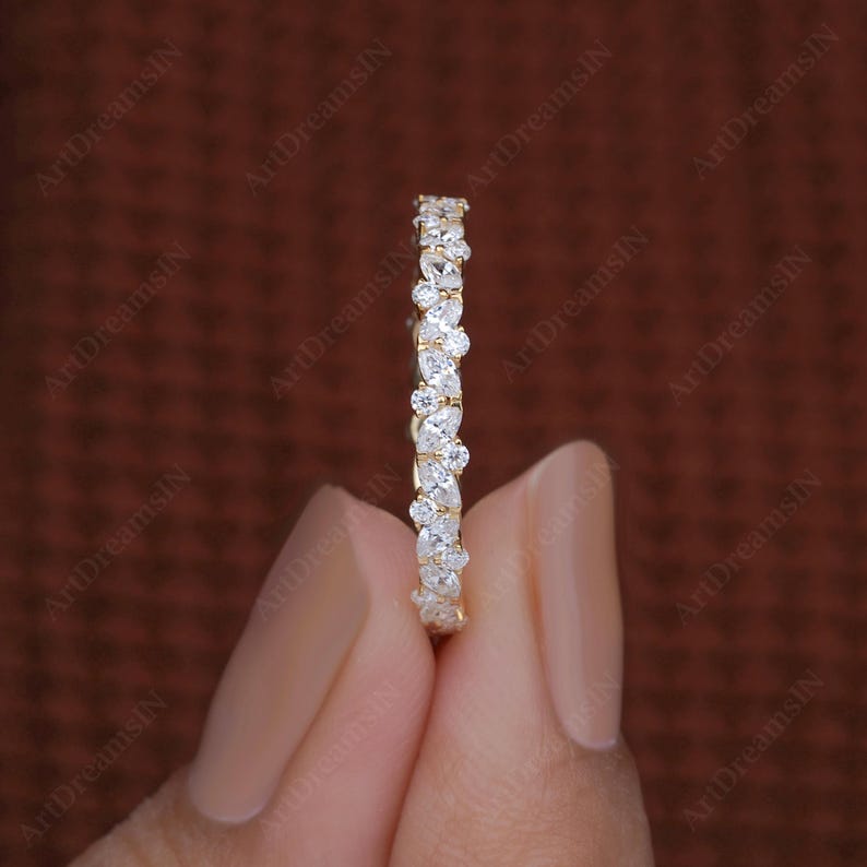 May include: A gold band ring with a continuous pattern of white gemstones. The ring is held by a person's hand against a dark brown background. The ring's gemstones are pear and round shaped.