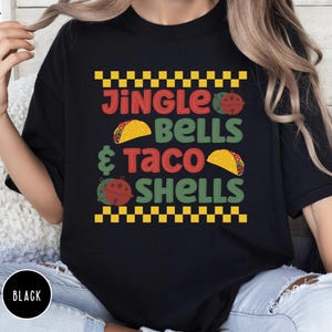 May include: Black t-shirt with a festive design featuring the text "Jingle Bells & Taco Shells" with a checkered pattern and images of tacos and jingle bells.