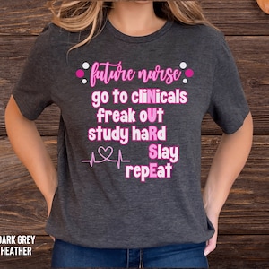 May include: Dark heather grey t-shirt with pink text that reads "future nurse go to clinicals freak out study hard slay repeat" with a pink heart and heartbeat graphic.