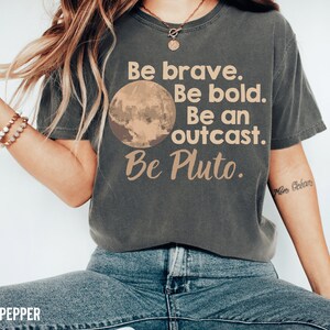 May include: A dark grey t-shirt with a graphic of a planet and the text "Be brave. Be bold. Be an outcast. Be Pluto."