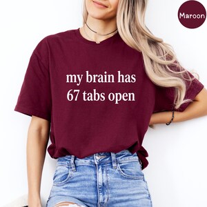 Könnte beinhalten: Burgunderfarbenes T-Shirt mit dem weißen Text "my brain has 67 tabs open". Das Shirt ist ein Rundhals-Stil und wird mit einer Jeans getragen. Das Wort "Maroon" befindet sich in einem Kreis in der oberen rechten Ecke.