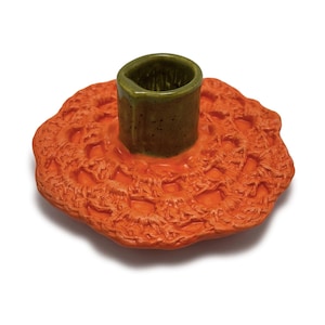 May include: A ceramic candle holder with a textured orange base and a cylindrical green center. The base has a detailed, organic pattern, and the center is a darker green. The candle holder is designed to hold a single candle.
