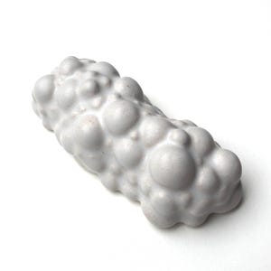 May include: A white, oblong sculpture with a textured surface of rounded, bubble-like shapes. The art piece is a solid, monochromatic form, with a matte finish. The object is set against a plain white background.