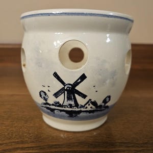 May include: White ceramic pot with a blue and white windmill design. The pot has three circular holes and a blue rim. The windmill scene is painted in blue and depicts a windmill, trees, and a small house.