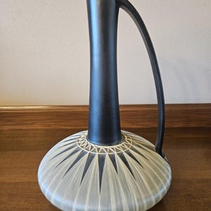 May include: A black ceramic vase with a wide, rounded base and a tall, slender neck. The base features a geometric pattern of white lines radiating outward from the center.