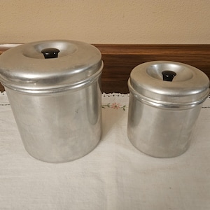 May include: Two vintage aluminium canisters with black knobs on a white tablecloth. The canisters are cylindrical with a slightly rounded top. The larger canister is approximately 18 cm tall, and the smaller one is about 13 cm tall.