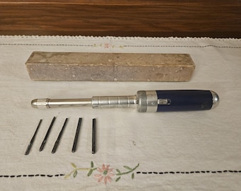 May include: A vintage screwdriver set with a dark blue handle and silver metal shaft. The set includes a rectangular cardboard box and several small, black screwdriver bits. The items are arranged on a white cloth.