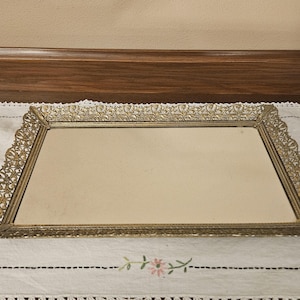 May include: A gold-colored metal tray with an ornate design. The tray has a rectangular shape and a mirror-like surface.