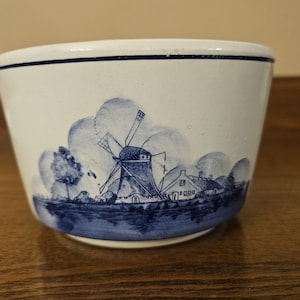 May include: White ceramic bowl with a blue windmill and landscape design. The bowl features a blue rim and a detailed illustration of a windmill, trees, and houses. The design is reminiscent of Delftware.