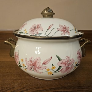 May include: White enamelware cooking pot with a lid, featuring pink lily floral designs and gold-colored accents. The pot has two handles and a gold-colored knob on the lid. A decorative kitchen item.