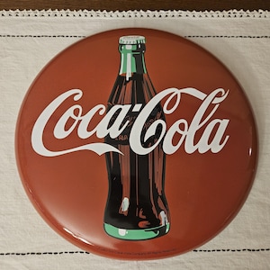 May include: A round, red Coca-Cola sign with a white Coca-Cola logo and a bottle of Coca-Cola in the center. The sign has a white border and the text "© 1963 The Coca-Cola Company. All Rights Reserved." at the bottom.