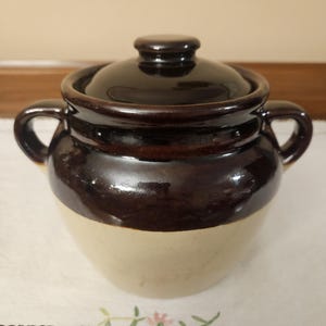 May include: A two-tone ceramic jar with a lid. The upper portion and lid are a dark brown, while the lower portion is a cream color. The jar has two small handles and is likely used for storage. The jar is sitting on a white surface.