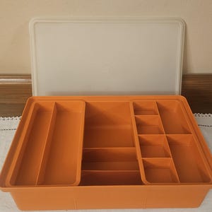May include: An orange plastic storage container with a white lid. The container has multiple compartments of varying sizes, ideal for organizing items. The lid is rectangular and translucent. The container is sitting on a white surface.