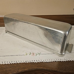 May include: A rectangular, aluminum loaf pan with rounded corners and a handle on one end. The pan is sitting on a white tablecloth with floral embroidery and a lace trim. The pan is designed for baking bread or cakes.
