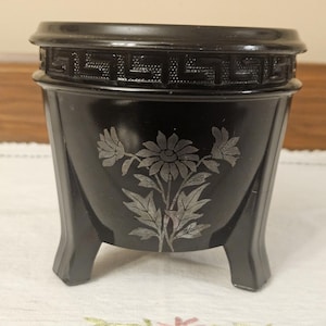 May include: A black, vintage-style container with a floral design. The container has a decorative band around the top and rests on three small feet. The floral design is etched into the front of the container.