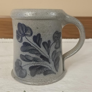 May include: A stoneware mug with a gray speckled finish and a dark blue floral design. The mug has a rounded handle and a slightly flared rim. The floral pattern is hand-painted and adds a rustic charm to the mug.