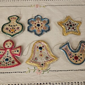May include: Six vintage Christmas ornaments made of paper mache and covered in metallic foil. The ornaments are decorated with colorful paint and glitter. The ornaments include a star, a bell, a bird, an angel, a snowflake, and a vase.