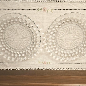 May include: Two clear glass plates with a textured, bubble-like pattern around the edges and a sunburst design in the centre. The plates are displayed on a white tablecloth with floral embroidery.