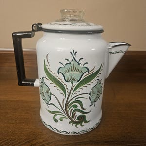 May include: White enamel coffee pot with a black handle and spout. The pot features a floral design in shades of green and brown. The lid has a clear glass knob. The pot is likely vintage and suitable for kitchen decor.