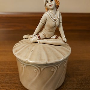 May include: A ceramic trinket box with a lid featuring a figurine of a woman in a swimsuit. The woman has red hair and is sitting on a scalloped edge. The box is decorated with a swirl pattern.