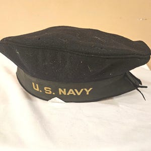 May include: A black wool U.S. Navy hat with a gold embroidered emblem.