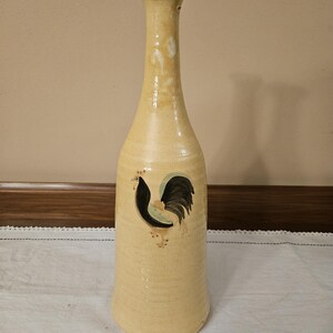 May include: A tall, yellow ceramic vase with a rooster design painted on the front. The rooster is black, green, and brown.