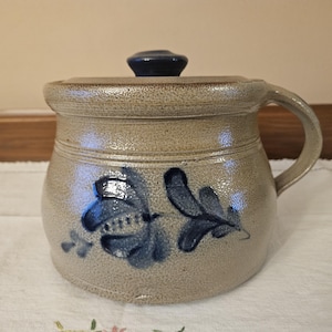 May include: A tan stoneware crock with a lid and handle. The crock features a blue floral design and a dark blue knob on the lid. The handle is curved, and the crock sits on a white surface.